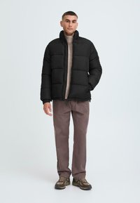 Svart pufferjakke med horisontale sømmer, høy krage og glidelås; kombinert med en ribbestrikket beige genser og brune tapered bukser.