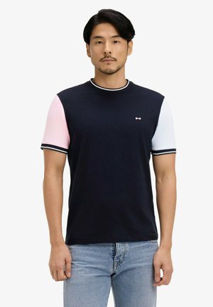Homme portant un T-shirt bleu marine avec une manche gauche rose, une manche droite blanche, un col et des poignets rayés, associé à un jean bleu clair.