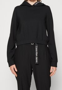 Mujer con sudadera negra con capucha con el logo de Calvin Klein en la capucha, combinada con pantalones negros que tienen un pasador de cinturón con la marca Calvin Klein Jeans.