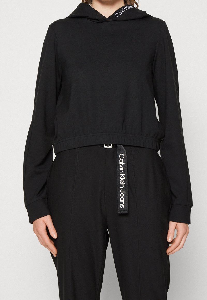 Mujer con sudadera negra con capucha con el logo de Calvin Klein en la capucha, combinada con pantalones negros que tienen un pasador de cinturón con la marca Calvin Klein Jeans.