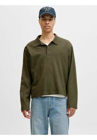 Polo-shirt à manches longues vert olive avec col, tissu texturé et patte de boutonnage à trois boutons, porté par-dessus une chemise blanche et assorti à un jean en denim clair.