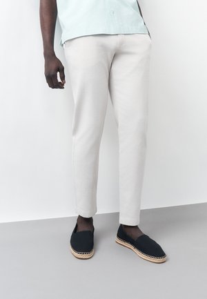 Férfi világos bézs slim fit nadrágban, világoskék ingben, fekete, szövött talpú espadrill csizmában, egyszínű fehér háttér előtt áll.