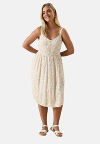 Kwiatowa sukienka midi w jasnym kremowym kolorze, z małymi różowymi i zielonymi kwiatami, regulowane ramiączka, dopasowana góra i marszczona talia. Nosi z białymi sandałami.