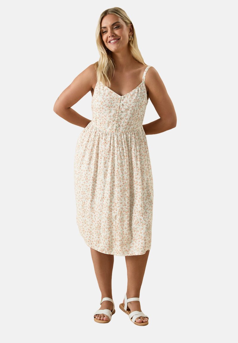 Kwiatowa sukienka midi w jasnym kremowym kolorze, z małymi różowymi i zielonymi kwiatami, regulowane ramiączka, dopasowana góra i marszczona talia. Nosi z białymi sandałami.