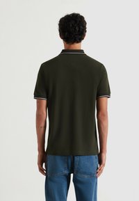 Polo shirt verde scuro con una texture sottile, dettagli bianchi sul colletto e sulle maniche, abbinato a jeans in denim blu chiaro.