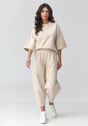 Kobieta z długimi falującymi włosami idzie, ubrana w luźną beżową bluzkę z krótkim rękawem i pasujące szerokie spodnie, na nogach białe sneakersy na platformie.