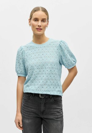 Frau trägt ein hellblaues Top mit Lochstickerei und Puffärmeln, kombiniert mit schwarzen, hoch taillierten Jeans und einem schwarzen Gürtel, steht mit der Hand in der Hüfte.