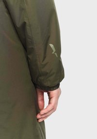 Veste olive avec une finition lisse et mate. Elle possède un poignet et un logo subtil de la même couleur sur la manche.