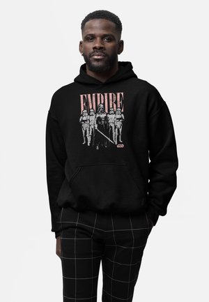Mężczyzna w czarnej bluzie z kapturem z grafiką Star Wars Empire z Darthem Vaderem i szturmowcami, w połączeniu z czarnymi spodniami w kratę.