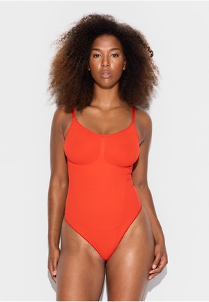 Žena s objemnými kučeravými vlasmi v priliehavých jasne oranžových jednodielnych plavkách stojaca na svetlom jednofarebnom pozadí.