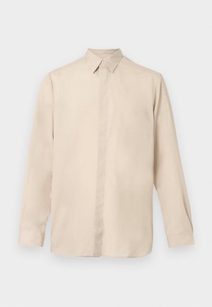 Beżowa koszula z długim rękawem wykonana z gładkiego materiału, z kołnierzem na guziki, pełną plisą z przodu i prostym dołem. Zaokrąglone mankiety.