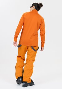 Oranges Langarm-Oberteil mit hohem Kragen, kombiniert mit orangen Schneehosen. Die Hosen haben kontrastierende graue Akzente und Taschen. Schwarze Schneestiefel sind sichtbar.
