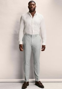 Weißes Langarmhemd mit Knöpfen, kombiniert mit hellgrauen taillierten Hosen. Braune Leder-Schuhanzüge vervollständigen den Look. Glatte Stofftextur.