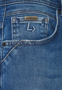 Dżinsy o średnim niebieskim odcieniu, z metalową metką na tylnej kieszeni, widocznym szwem i subtelnym logo w kształcie litery "L".