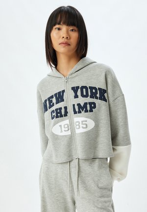 Grijze korte rits-hoodie met "NEW YORK CHAMP 1985" in donkerblauw en wit gedrukt. Heeft een kap en contrasterende mouwen.
