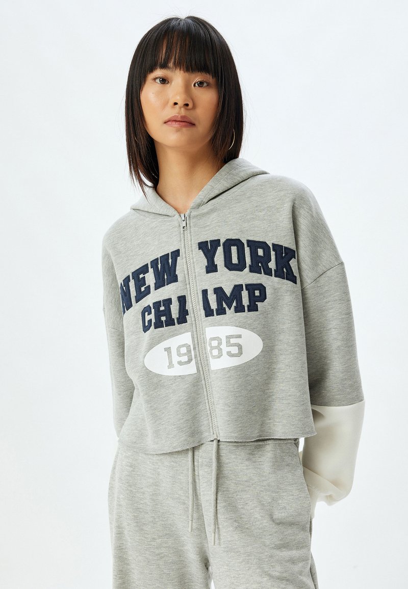 Felpa corta grigia con zip e stampa "NEW YORK CHAMP 1985" in blu scuro e bianco. Presenta un cappuccio e polsini delle maniche a contrasto.