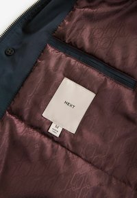 Jachetă bleumarin cu fermoar, cu căptușeală bordeaux și un model subtil. Include o etichetă cu "NEXT" și mărimea "M" pe un tag din țesătură.