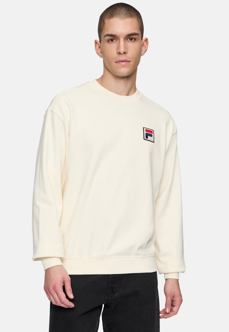 Sudadera color crema con mangas largas, puños y dobladillo de canalé; presenta un pequeño logotipo bordado en rojo, blanco y azul en el pecho.