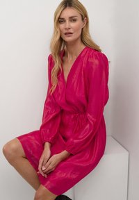 Femeie cu păr lung blond, așezată, purtând o rochie lungă cu mâneci lungi, lucioasă și roz aprins, cu talie strânsă, pe un fundal alb simplu.