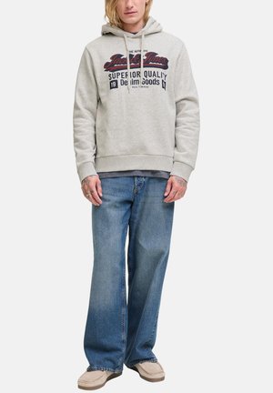 Persona che indossa un hoodie grigio chiaro con logo, jeans blu larghi e scarpe beige, in piedi contro uno sfondo bianco uniforme.