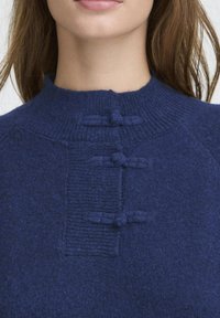 Tums zila vilnas džemperis ar augstu apkakli un faktūru bagātu priekšpusi, kas ir izrotāta ar trim dekoratīvām mezglēm un ribotu detaļu.