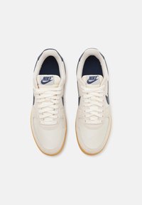 Nike tenisky s krémovým plátěným svrškem, námořnickými akcenty, bílými šňůrkami a gumovou podrážkou. Kulatá špička a polstrované podšívka pro pohodlí.