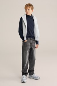 Grå sweatshirt som bärs över en marinblå långärmad skjorta, ihop med mörkgrå jeans och silverfärgade träningsskor, mot en neutral bakgrund.