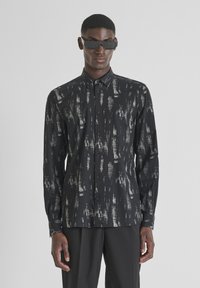 Zwart overhemd met lange mouwen en een zilveren abstract patroon, met knoopsluiting aan de voorkant en een kraag. Gedragen met vierkante zwarte zonnebril.