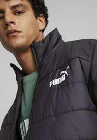 Giacca nera isolata con pannelli cuciti, colletto alto e zip frontale. Presenta un logo Puma bianco ben visibile sul lato sinistro del petto.