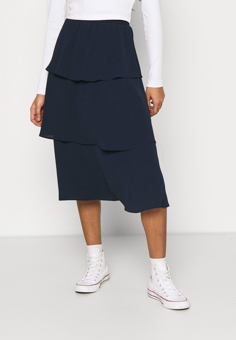 Femme portant une jupe midi à volants bleu marine, un haut à manches longues blanc, des chaussettes blanches et des baskets montantes blanches, se tenant sur un fond uni.