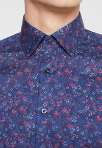 Koszula w ciemnym niebieskim kolorze z kwiatowym wzorem, z małymi czerwonymi i jasnoniebieskimi kwiatami. Kołnierzyk z dopasowanymi guzikami. Materiał o teksturze.
