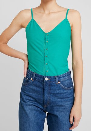 Kvinde iført teal ærmeløs top med knapdetaljer kombineret med blå højtsiddende jeans, hånd hvilende på hoften mod ensfarvet baggrund.