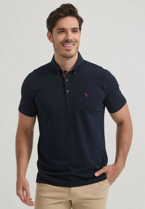 Homme souriant aux cheveux foncés portant un polo bleu marine avec un logo rouge et un pantalon beige, debout avec une main dans la poche.