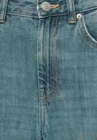 Gros plan sur un jean en denim bleu montrant le bouton, la zone de la fermeture éclair, les passants de ceinture et une partie de la poche avant avec des coutures visibles.