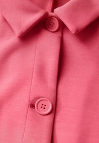 Rosa polo-tröja med en slät tygstruktur, med krage och två knappar. Knapparna har rund form med fyra hål.