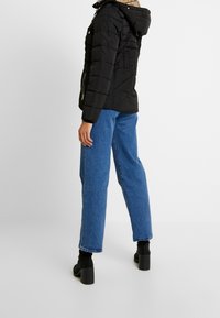 Giubbotto imbottito nero con cappuccio, dotato di una superficie testurizzata e tasche con zip. Indossato con jeans blu e stivaletti neri con tacco.