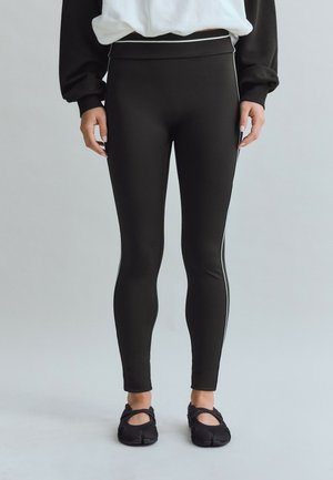 Person iført sorte leggings med hvide sidelommer og sorte split-toe sko, stående mod en ensfarvet lys baggrund.