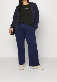Cárdigan de punto azul marino sobre una camiseta negra con el logo blanco de "TOMMY JEANS", combinado con pantalones azul marino y zapatillas beige.