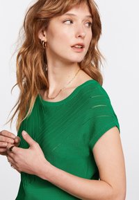 Grön stickad topp med ett texturerat mönster med diagonala ränder, korta ärmar och en bred rund halsringning. Lätt och figurnära design.