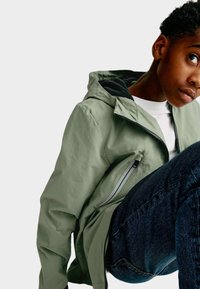Giacca verde chiaro con cappuccio, fodera nera e dettagli con zip, abbinata a jeans blu scuro. Tessuto liscio con una vestibilità casual.