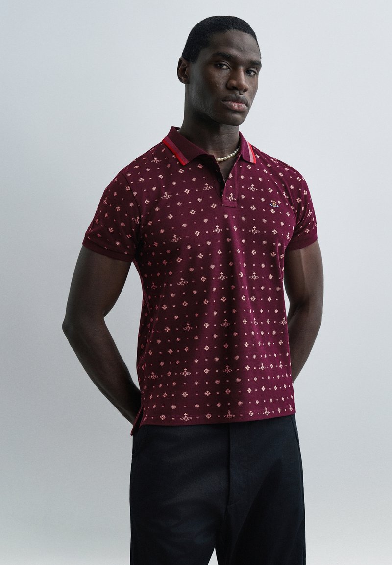 Άνδρας που φοράει πουκάμισο πόλο μ ε μοτίβο σε μπορντό χρώμα, μαύρο παντελόνι και κολιέ από μαργαριτάρια, όρθιος με τα χέρια πίσω από την πλάτη, μπροστά σε ένα απλό φόντο.