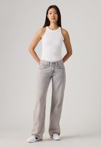 Λευκό ριγωτό αμάνικο τοπ σε συνδυασμό με ανοιχτόχρωμα τζιν παντελόνια με φαρδιά πόδια. Το μοντέλο φορά λευκά αθλητικά παπούτσια, με τα χέρια στη μέση, ουδέτερο φόντο.