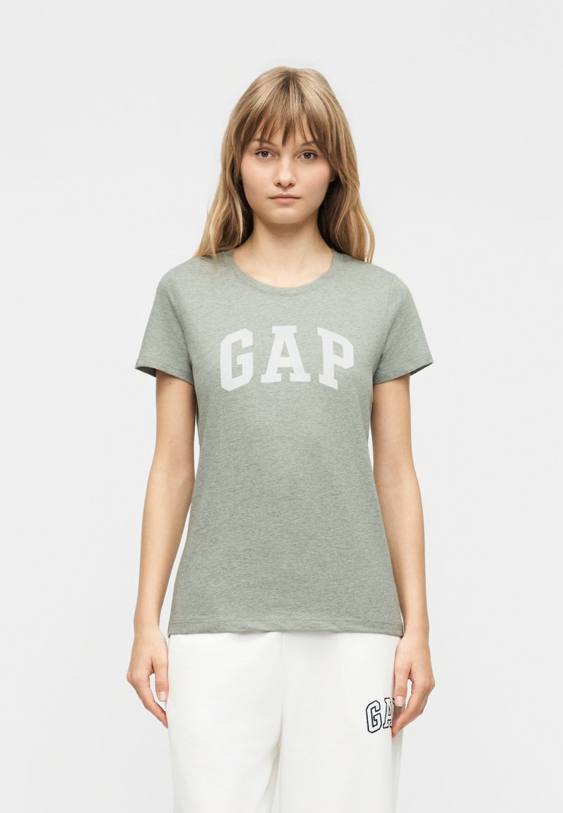Jeune femme aux cheveux châtain clair portant un T-shirt gris GAP et un pantalon blanc avec un logo GAP sur la cuisse gauche, debout devant un fond uni.