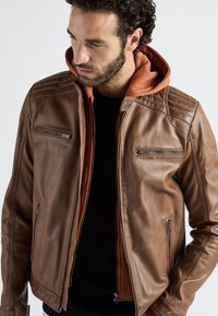 Bruine leren jas met een ritssluiting aan de voorkant, twee borstzakken en een oranje capuchon. Soepele textuur met gewatteerde schouderdetails.