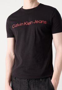 Černé bavlněné tričko s krátkým rukávem, s moderním červeným logem "Calvin Klein Jeans" přes hruď.