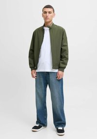 Veste bomber olive avec fermeture éclair, t-shirt à col rond blanc, jean large bleu clair, et baskets noires avec accents blancs.