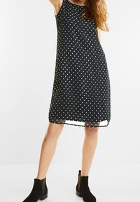 Svart ärmlös klänning med vita prickar, rak silhuett och en genomskinlig fåll. I kombination med svarta ankelstövlar.