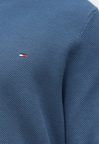 Zils teksturēts adījuma audums ar mazu sarkanu, baltu un melnu taisnstūra logotipu, kas ir izsistīts tuvu centrālajai kreisajai daļai.