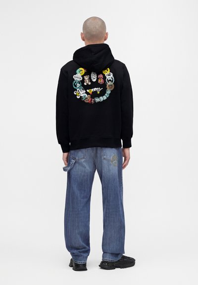 Homme portant un sweat à capuche noir avec des patchs colorés en forme de smiley dans le dos, associé à un jean bleu ample et des baskets noires, de dos.