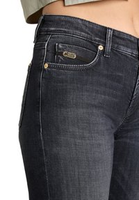 Dunkelblaue Denim-Jeans mit schmaler Passform, goldfarbenen Beschlägen und einer kleinen vorderen Tasche mit einem metallischen Markenzeichen. Glatte Textur sichtbar.